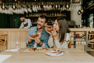 Neşeli bir çift kırsal bir restoranda bir anı paylaşıyor, pizza dilimlerinin tadını çıkarırken birlikte gülüyorlar ve çevrelerindeki canlı atmosferin tadını çıkarıyorlar..
