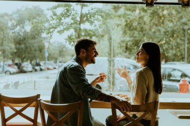 İki birey, arka planda ağaçlar ve arabalarla büyüleyici bir kafede lezzetli bir yemeğin tadını çıkarırken kahkahalarını ve iç içe geçen sohbetlerini paylaşıyor..