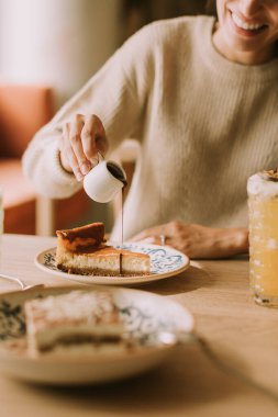 Büyüleyici bir kafe ortamında, gülümseyen bir kadın bir dilim peynirli kekin üzerine ustalıkla şurup serpiyor, sıcaklığın ve huzurun tadını çıkarıyor..