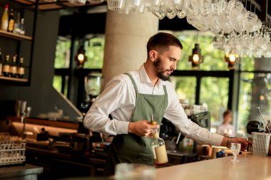 Barmen, doğal ışıkla dolu modern bir barda bir dizi cam eşyayla çevrili bir kokteyli ustalıkla hazırlarken konsantre oluyor ve davetkar bir atmosfer yaratıyor.