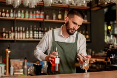 Canlı bir bar ortamında, kendine güvenen bir barmen yeni bir kokteyl hazırlar, uzmanlığını elinde bir şişeyle gösterir, gecenin telaşına hazırdır.