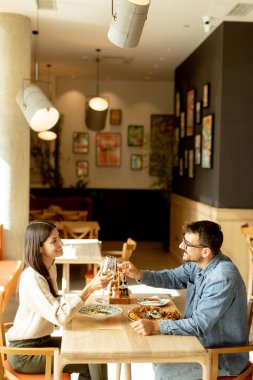 Bir çift kadehlerini neşeli bir şekilde kaldırır, ahşap bir masada birbirlerine gülümserler, canlı tabaklarla süslenirler, sıcak ve samimi bir atmosfer yaratırlar..