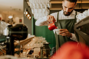 Sıcak ve davetkar bir kafede, kendini adamış bir barista kahve bardağına ustalıkla süt dökerek müşteriler için hoş bir deneyim yaratıyor..