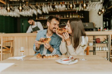 Neşeli bir çift kırsal bir restoranda bir anı paylaşıyor, pizza dilimlerinin tadını çıkarırken birlikte gülüyorlar ve çevrelerindeki canlı atmosferin tadını çıkarıyorlar..
