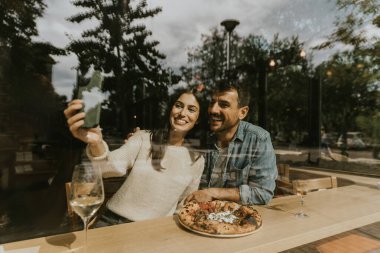 Bir çift, ahşap bir barda oturur, kahkahalar atar ve keyifli bir anı paylaşırlar. Önlerinde lezzetli bir pizzayla selfie çekerken, güneşin altında güneşlenirken..
