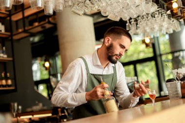 Barmen, doğal ışıkla dolu modern bir barda bir dizi cam eşyayla çevrili bir kokteyli ustalıkla hazırlarken konsantre oluyor ve davetkar bir atmosfer yaratıyor.