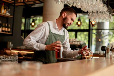 Canlı bir bar ortamında, kendine güvenen bir barmen yeni bir kokteyl hazırlar, uzmanlığını elinde bir şişeyle gösterir, gecenin telaşına hazırdır.