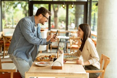 Neşeli bir çift, güzel bir kafe masasında oturur, güzel bir yemek ve güneşli bir atmosferle çevrili akıllı bir telefondan anı yakalarken gülümserler.