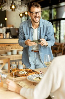 Neşeli bir çift, güzel bir kafe masasında oturur, güzel bir yemek ve güneşli bir atmosferle çevrili akıllı bir telefondan anı yakalarken gülümserler.
