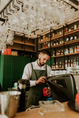 Şık bir kafe içinde, yetenekli bir barista, düzenli düzenlenmiş cam süslemeler ve canlı dekorlarla çevrili lezzetli içecekler hazırlamaya odaklanıyor, müşteriler için sıcak bir ortam yaratıyor..