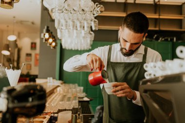 Sıcak ve davetkar bir kafede, kendini adamış bir barista kahve bardağına ustalıkla süt dökerek müşteriler için hoş bir deneyim yaratıyor..