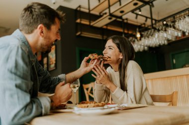 Neşeli bir çift kırsal bir restoranda bir anı paylaşıyor, pizza dilimlerinin tadını çıkarırken birlikte gülüyorlar ve çevrelerindeki canlı atmosferin tadını çıkarıyorlar..