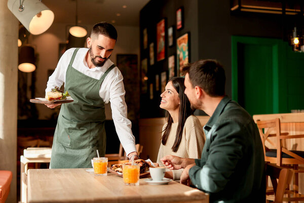 In a lively cafe filled with colorful artwork, a waiter presents fresh vegetarian meals to a smiling couple enjoying their afternoon together over refreshing drinks