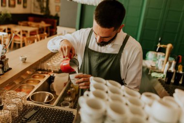 Sıcak ve davetkar bir kafede, kendini adamış bir barista kahve bardağına ustalıkla süt dökerek müşteriler için hoş bir deneyim yaratıyor..