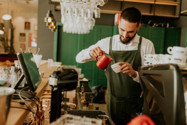 Sıcak ve davetkar bir kafede, kendini adamış bir barista kahve bardağına ustalıkla süt dökerek müşteriler için hoş bir deneyim yaratıyor..