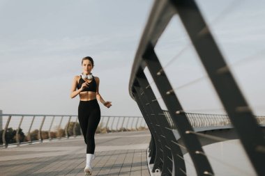 Siyah atletik kıyafetli fit bir kadın gösterişli bir köprüde koşuyor ve akşam rutininin tadını çıkarıyor. Ufuk hafifçe parlıyor, kararlılığını ve uygunluğa bağlılığını vurguluyor.