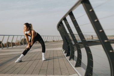 Derin bir nefes alan bir kadın, vücudunu egzersize hazırlar. Pürüzsüz bir köprüde uzanarak, sabahın erken saatlerinde suyun yanında sakinliğin tadını çıkararak.