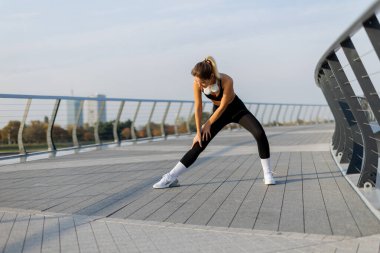 Derin bir nefes alan bir kadın, vücudunu egzersize hazırlar. Pürüzsüz bir köprüde uzanarak, sabahın erken saatlerinde suyun yanında sakinliğin tadını çıkararak.