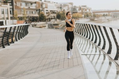 Siyah atletik kıyafetli fit bir kadın gösterişli bir köprüde koşuyor ve akşam rutininin tadını çıkarıyor. Ufuk hafifçe parlıyor, kararlılığını ve uygunluğa bağlılığını vurguluyor.
