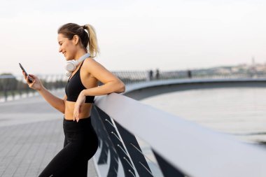 Bir kadın, gün batımında nehrin kenarındaki huzurlu ortamın tadını çıkarırken, kulaklıkla müzik dinlemekten zevk alır..