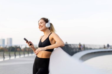 Genç bir kadın, bir köprüde, kulaklıkları takmış, günbatımının hafif parıltısında suyun kenarında günbatımının tadını çıkarırken müziğin tadını çıkarıyor..