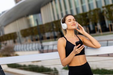 Çağdaş bir binanın dışında, bir kadın, şehir parkının güneşli atmosferinin tadını çıkarırken kulaklıkla müzik eşliğinde çalışmaktan zevk alır.