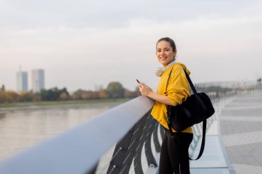 Kendine güvenen bir gülümsemesi olan genç bir kadın, parlak sarı kapüşonlu ve taytlı bir öğleden sonra için rahat giyinmiş, nehir kenarındaki cam korkuluklarda geziniyor..