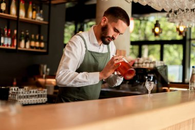 Canlı bir bar ortamında, kendine güvenen bir barmen yeni bir kokteyl hazırlar, uzmanlığını elinde bir şişeyle gösterir, gecenin telaşına hazırdır.