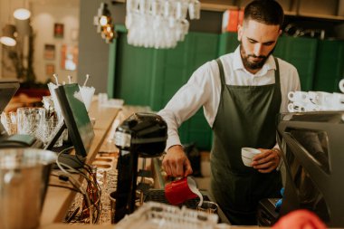 Sıcak ve davetkar bir kafede, kendini adamış bir barista kahve bardağına ustalıkla süt dökerek müşteriler için hoş bir deneyim yaratıyor..