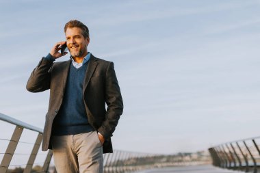 Akıllı takım elbiseli bir adam, parlak mavi bir gökyüzünün altında, parlak, çağdaş bir köprü boyunca rahatça dolaşır, bir telefon görüşmesi yapar..