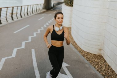 Sportif kıyafetler giymiş bir kadın, su kenarında bir patika boyunca mutlu bir şekilde koşar. Kulaklıklarıyla, canlı sabah atmosferini ve serin esintiyi kucaklıyor..