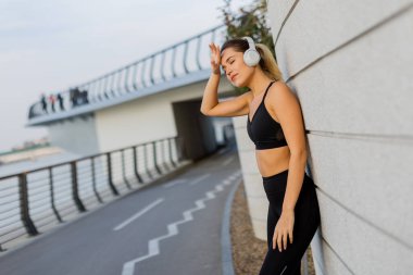 Sporcu genç bir kadın şık bir duvara yaslanmış, kulaklık takmış, enerjik bir etkinlikle dolu canlı bir şehir ortamında koştuktan sonra müziğinin tadını çıkarıyor..
