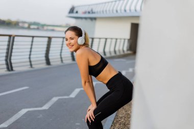 Yoğun bir koşudan sonra, genç bir kadın nefes almak için durur, canlı bir şehir ortamında kulaklıklarıyla müzik dinlerken gülümseyerek..