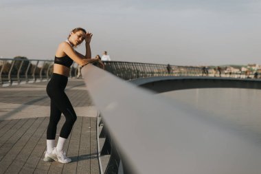 Atletik giyinmiş genç bir kadın zarif bir korkuluğa yaslanıyor, gücünü ve zarafetini sergiliyor ve nehrin kıyısında başkalarıyla akşam yürüyüşünün keyfini çıkarıyor..