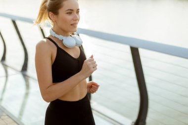 Atletik giyinmiş genç bir kadın nehir kenarında enerjik bir şekilde koşuyor, güzel bir sabahın dingin atmosferini kucaklıyor, en sevdiği müzikle besleniyor..