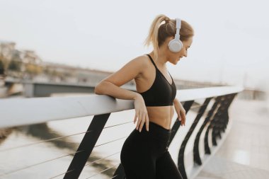 Spor kıyafetleri giymiş genç bir kadın müzik dinlemek için koşuya ara veriyor. Huzurlu bir arka plan ve şehir hayatı olan korkuluklara yaslanıyor..