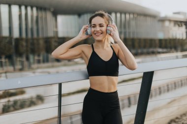 Atletik olarak gülümseyen bir kadın kulaklıkla müzik dinler. Güneş batarken korkuluklardan mutluluk ve enerji saçar..