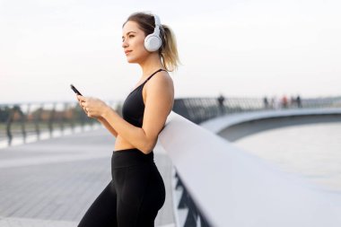 Genç bir kadın, bir köprüde, kulaklıkları takmış, günbatımının hafif parıltısında suyun kenarında günbatımının tadını çıkarırken müziğin tadını çıkarıyor..