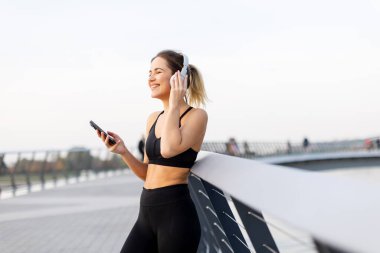 Genç bir kadın, bir köprüde, kulaklıkları takmış, günbatımının hafif parıltısında suyun kenarında günbatımının tadını çıkarırken müziğin tadını çıkarıyor..