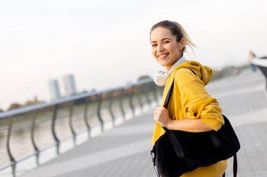 Neşeli bir kadın, parlak sarı bir kapüşonlu ve kulaklıklı, güzel bir rıhtım yolunda yürüyor. Onun canlı gülümsemesi, günün güneşli atmosferini yansıtır..