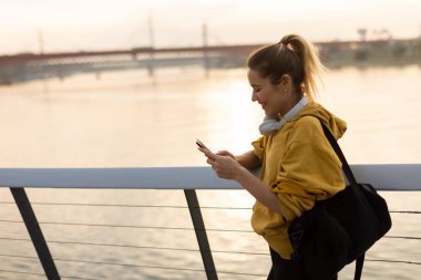 Suyun üzerinde güneş batarken, sarı kapüşonlu genç bir kadın nehir kenarında duruyor, kulaklık takmış, sükuneti kucaklıyor..