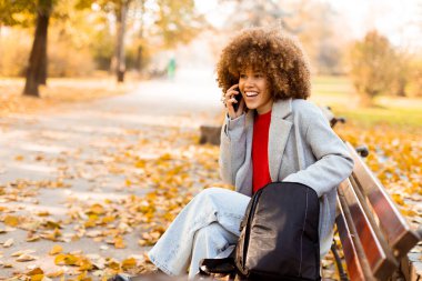 Kıvırcık saçlı neşeli bir kadın, canlı bir sonbahar yeşillikleriyle çevrili bir bankta oturmuş, dışarıda güzel bir günün tadını çıkarırken telefon eder..