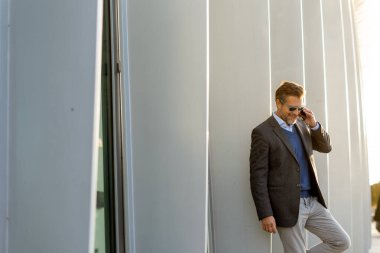 İyi giyimli bir adam telefonuyla konuşurken parlak beyaz bir duvara yaslanıyor. Günbatımının sıcacık ışığı etrafındaki modern mimariyi güzel bir şekilde vurgular..