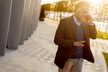 İyi giyimli bir adam, çağdaş binaların yanında güneşli bir yolda yürürken telefonda sohbet etmekten zevk alır ve sofistike bir ortam yaratır..