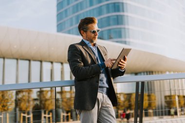 Şık bir adam bir balkonda duruyor, tabletle gösterişli bir mimarinin arka planına saldırıyor..