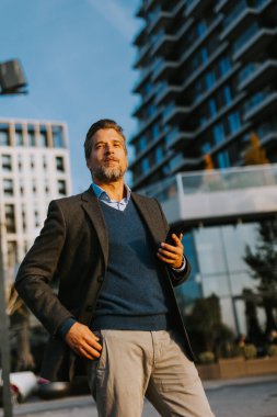 Ceket ve süveter giymiş neşeli bir adam dışarıda duruyor, akıllı telefonunu canlı şehir atmosferine ve açık mavi gökyüzüne dalmış, hoş bir gülümsemeyle tutuyor..