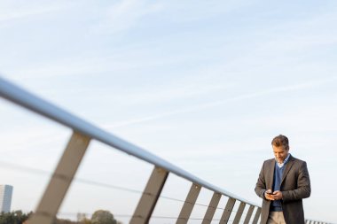 Resmi bir takım elbise giymiş bir işadamı, güneş batarken akıllı telefonuna odaklanmış şık bir köprüde duruyor. Şehrin ufuk çizgisi çarpıcı bir zemin oluşturuyor..
