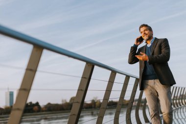 Akıllı takım elbiseli bir adam, parlak mavi bir gökyüzünün altında, parlak, çağdaş bir köprü boyunca rahatça dolaşır, bir telefon görüşmesi yapar..