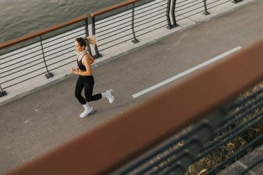 Güneş sakin suyun üzerinde yükselirken, kararlı genç bir kadın güzel bir sahil yolunda koşar, doğanın içinde zindelik ve refahını gösterir..