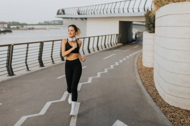 Sabahın erken saatlerinde, zinde genç bir kadın nehir kenarında zarif bir şekilde koşar, doğanın güzelliği ve çağdaş tasarım elementleriyle uyum sağlar..